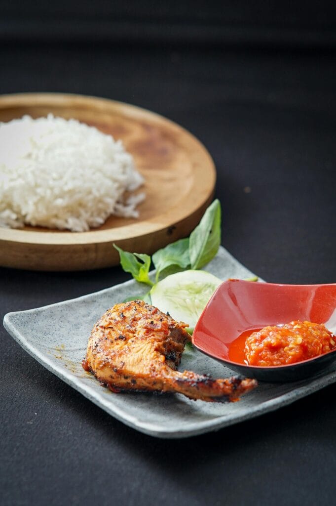 Grilled chicken served with spicy sambal, white rice, and cucumber slices on elegant plates.